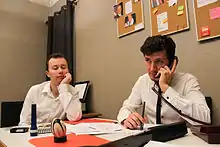 Deux personnages autour d'un bureau dans un bureau ; l'un, affalé, pensif, l'autre, au téléphone, sérieux.