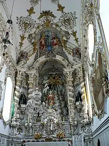 Photographie en couleurs. Dans un intérieur d'église très éclairé, un grand retable blanc avec des décorations dorées s'inscrit dans un cadre de la même configuration.