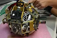 Un des satellites SPHERES sans sa coque en plastique. La structure en aluminium, le panneau de contrôle, les capteurs à ultrasons, propulseurs, la molette du régulateur de pression et le manomètre de pression sont visibles.