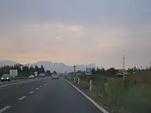 Photographie dans le sens longitudinal d'un axe routier traversant la campagne ; panneaux routiers ; véhicules qui se croisent sur deux voies doubles et en arrière plan un décor de montagnes cisaillées par le ciel.