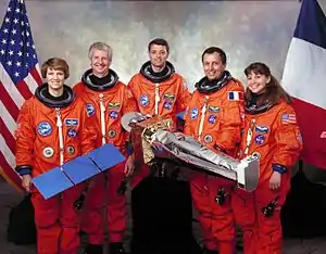 Deux femmes et trois hommes, en tenue d'astronaute, devant un drapeau américain et un drapeau français.