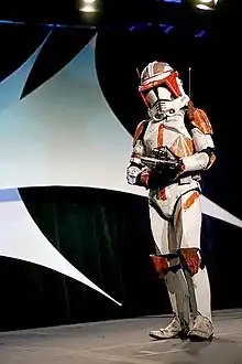 Homme portant une armure futuriste intégrale blanche, un fusil, avec un casque à visière décoré de orange.