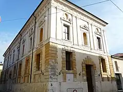 Vue d'en-dessous d'un bâtiment carré blanc ; au rez-de-chaussée les portes et fenêtres sont entourés de grosses pierres jaunes ; à l'étage les fenêtres sont décorées à l'aide de moulures aux formes classiques.