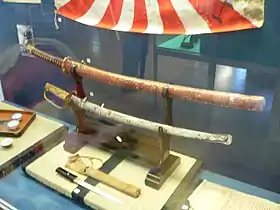 Pièces d'époque d'épées militaires japonaises reposant devant un a'tanto sur leur katana kake, de haut en bas : le modèle « nouveau » (shin guntō), 1935 – 1945 ; le modèle « ancien » (kyū guntō), ou Muratatō (1875 – 1934).
