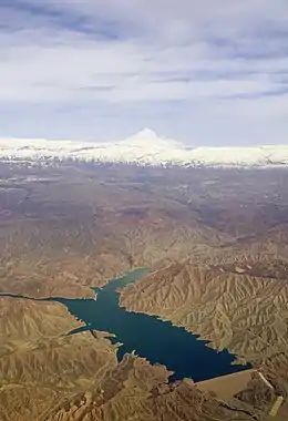 Vue aérienne un lac de retenue au bleu profond, des montagnes ocres dénuées de végétation, en arrière-plan des crêtes plus hautes et un cône volcaniques enneigés.