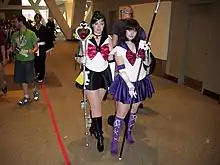 Deux jeunes filles grimées et déguisées en personnage de la série Sailor Moon. Sailor Pluto est à gauche de l’image, et a les cheveux longs et verts, porte une robe noire et rouge. Sailor Saturn est à droite, elle a les cheveux de couleur noire. Son costume est violet et elle possède une longue faux.