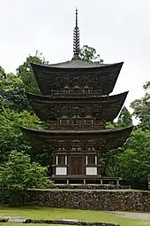 Photo couleur d'une pagode en bois de couleur marron installée sur un socle en pierre. En arrière-plan : des arbres au feuillage vert et un ciel blanc laiteux.
