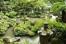 Photo couleur d'un jardin paysager japonais comprenant des massifs de verdure, des arbres, des rochers, un étang et, au premier plan, une lanterne de pierre (couleur dominante : vert).
