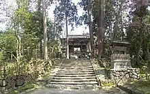 Photo couleur d'un escalier en pierre menant à la porte en bois à un étage d'un temple bouddhique. Des arbres de part et d'autre de l'escalier, un ciel blanc laiteux en arrière-plan.