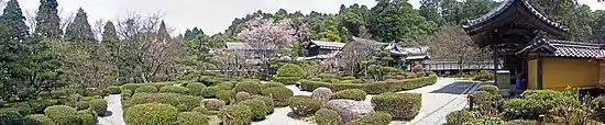 Photo couleur panoramique de l'intérieur d'un parc comprenant, au premier plan, des massifs feuillus sur un sol graveleux blanc, et des arbres en arrière-plan.