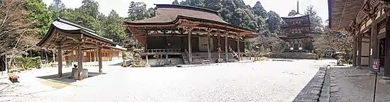 Photo couleur de l'enceinte d'un temple bouddhique avec, au centre, un hall en bois marron à un étage, au fond, une pagode à trois étages, à gauche, un pavillon d'ablution, et, au premier plan, un sol graveleux gris clair. En arrière-plan : des arbres au feuillage vert.