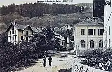 Carte postale, photographie de l'entrée du village : habitations à gauche et à droite, deux passants de dos au milieu, les pâturages au fond.