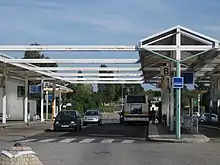 La photographie présente un centre bus en été. Un bus de la SEMTAO stationne sur le quai B, une personne descend du bus. Des voitures circulent sur le quai A. Le quai C est vide. Les préaux des quais sont soutenus par des piliers métalliques peints en blanc.