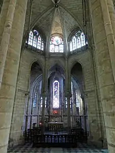 Chapelle contenant quelques chaises et présentant des verrières bleutées et noircies, surmontée de trois vitraux décoratifs et d'un vitrail figuré.