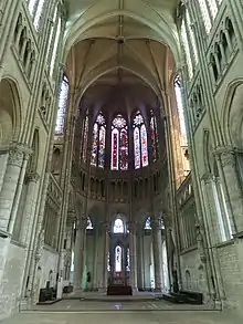 Intérieur d'une église avec des voûtes très hautes soutenues par des piliers en pierre. En haut de l'image, grandes verrières colorées. En bas de l'image, 5 ouvertures délimitées par 6 gros piliers donnent accès à des chapelles.