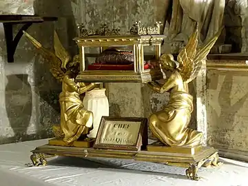 Deux anges féminins ailés et dorés tiennent une boite présentant des ouvertures vitrées sur les côtés et contenant un morceau de tissu posé sur un coussin.