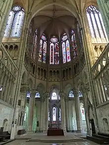Intérieur d'une église avec des voûtes très hautes soutenues par des piliers en pierre. En haut de l'image, grandes verrières colorées. En bas de l'image, 5 ouvertures délimitées par 6 gros piliers donnent accès à des chapelles.
