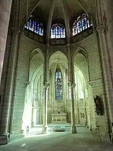 Chapelle contenant un gisant de pierre et une statue accrochée au mur de droite, surmontée de trois vitraux décoratifs.