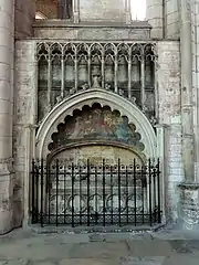 Tombeau incrusté dans un mur, séparé du déambulatoire par une grille en fer forgée, avec un gisant de pierre surmonté par une peinture murale représentant un homme auréolé entouré de deux anges.