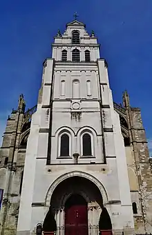 Tour-porche présentant une couleur très blanche. En haut de la tour, une croix surmontée d'un coq dorée et, un peu plus bas, deux pots à feu. Ciel bleu.