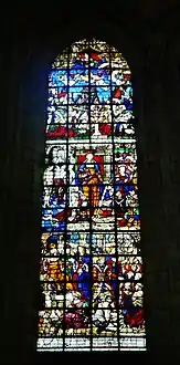 Grand vitrail représentant une vingtaine de personnages. Au centre de l'image, une femme habillée en orange porte une épée et un livre rouge. Des hommes sont agenouillés à ses pieds.