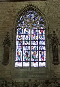 Au centre de l'image, vitrail légèrement bleutée représentant quatre femmes en robe. De part et d'autre du vitrail, des niches sont incluses dans un mur de pierre. Dans l'une des niches, une statue d'homme barbue est conservée.