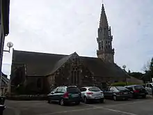 Au premier plan la place de l'église et ses voitures garées, au second plan l'église avec son clocher à droite.