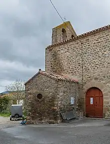 Vue de l'église Saint-Marc : des murs en pierres apparentes, une porte voutée rougeâtre, on devine le mur-clôcher par dessus le toit en tuile. Le ciel est nuageux.