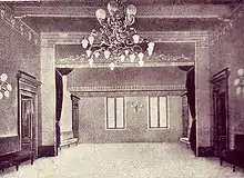 Photographie en noir et blanc d'une salle vide, en perspective : deux portes à gauche et deux portes à droite séparées par des banquettes, deux fenêtres sur le mur du fond, frises au plafond, lustre et appliques murales.