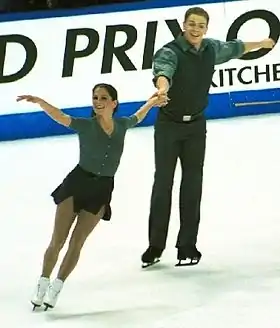 Photographie d'un couple de patineurs en compétition, se tenant par la main, la femme devant l'homme.
