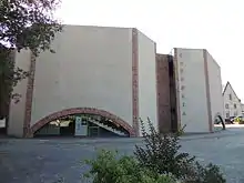 Photographie d'un bâtiment aux hauts murs blancs aux arêtes de brique. Sur la façade visible à gauche, un arc de brique, surbaissé et vitré, laisse voir un escalier. Sur un mur à droite, les lettres COPPELIA, verticales, en majuscules.