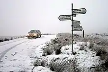 Photographie couleur de Sally Gap sous la neige avec une voiture, phares allumés, passant au niveau d'un panneau indiquant plusieurs directions et distances
