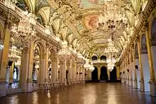 Vue d'un salon de l'hôtel de ville de Paris. La pièce est vide, on distingue le parquet, les lustres pendus au plafond et les moulures dorées.