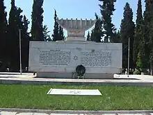 Un mémorial de pierre blanche surmonté d'une menorah, en arrière fonds des cyprès.