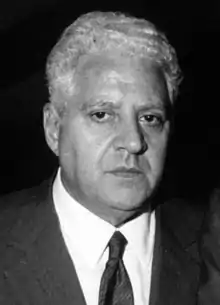 Image en noir et blanc du visage d'un homme, cheveux blancs, chemise blanche et cravate, regardant le spectateur.