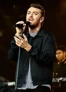 Photographie couleur d'un homme qui chante dans un micro et lève le bras gauche.