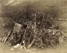 Vue d'un groupe de personnes devant une hutte constituée de pierres et de branches d'abres