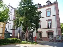 Bâtiment à deux étages crépi rosé très clair avec des parements de grès rose. Au premier plan, un grand arbre aussi haut que le bâtiment.