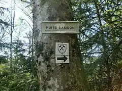 Panneau portant la mention « puits Samson » et une flèche dirigée vers la gauche tandis que le sentier minier est indiqué vers la droite.