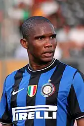 Photo en buste et de trois-quart d'un footballeur noir portant un maillot avec des rayures verticales noires et bleues