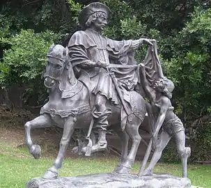 Photographie en couleurs d'une statue en bronze : un cavalier coupe une partie de son vêtement et la donne à un homme à pied.
