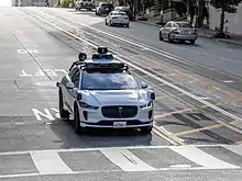 Une voiture Waymo blanche arrêtée devant un passage piéton dans une rue de San Francisco. Le véhicule est bardé de capteurs, en particuliers sur son toit où trois niveaux de capteurs scannent les alentours.