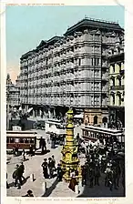 Photographie ancienne d'une rue agrémentée d'une fontaine et dans laquelle passent de nombreux usagers.