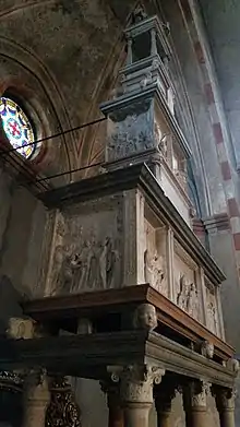 Image d'une vue de profil d'un sarcophage en marbre décoré de reliefs posé sur des colonnes de candélabres soutenu par des têtes de putti, avec un fronton en forme de dôme surmonté d'un édicule.