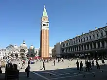 Image photographique d'une place avec un grand campanile en brique rouge et sommet blanc, sur la gauche un grand palais blanc à portiques et fenêtres superposées et sur le fond une basilique dont la façade se compose de cinq portails dont un central plus grand et le toit de trois dômes.