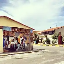 Photographie d'un rue de village avec sur la gauche une maison dont le mur est peint d'une scène de vie représentant des toits en tuiles, murs, rue et cinq personnages dans différentes positions