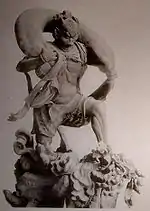 Fūjin. Three-quarter view of a statue. His left leg is bend as if climbing stairs et he is carrying a long bag-shaped object which goes from one shoulder to the other around the back of his head. Black and white photograph.
