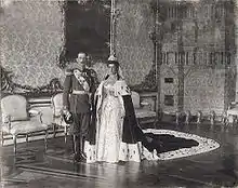Cliché noir et blanc dans grand salon. Jeune homme : uniforme militaire. Jeune femme en mariée et long manteau impérial.