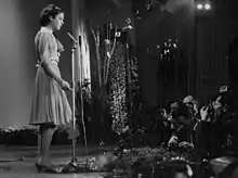 Photographie en noir et blanc, sur une scène, avec sur la gauche, une jeune fille en robe, chantant devant un micro, en bas, sur le coin droit, divers spectateurs