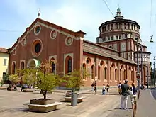 Image photographique d'un édifice religieux dont la façade simple à pignon est divisée en cinq éperons avec six contreforts avec un portail central en marbre blanc surmonté d'une lunette et au fond à droite une grande coupole.